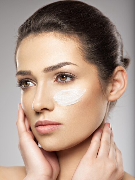 Hermoso rostro de mujer joven con frotis de crema cosmética en la cara cerca del ojo. Concepto de cuidado de la piel. Concepto de tratamiento de belleza.