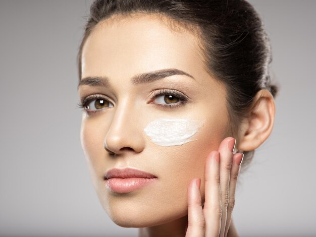 Hermoso rostro de mujer joven con frotis de crema cosmética en la cara cerca del ojo. Concepto de cuidado de la piel. Concepto de tratamiento de belleza.