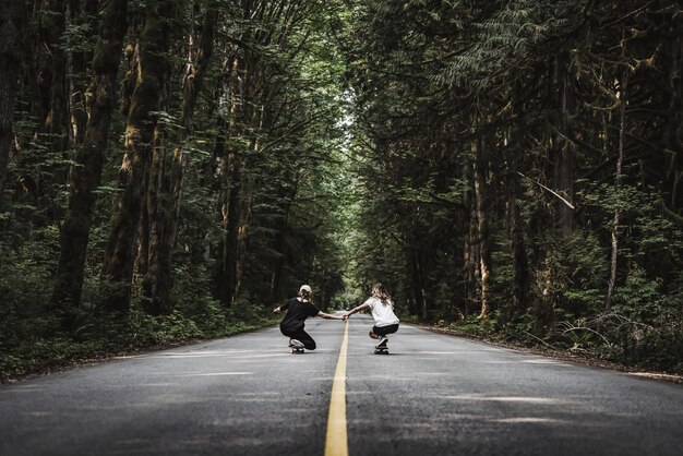 Hermoso tiro de hembras cogidos de la mano blanca patinando sobre una carretera vacía en medio del bosque