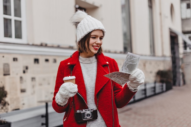 Hermoso turista con sombrero blanco y abrigo rojo con mapa, explorando la ciudad. Retrato de niña con guantes en el fondo del edificio.