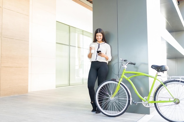 Hermoso viajero sonriente sosteniendo una taza desechable y un teléfono inteligente en bicicleta en el pasillo de la oficina