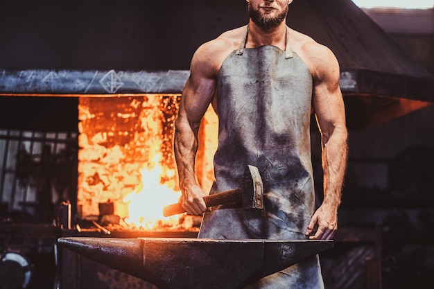 Herrero musculoso en delantal protector en su taller con martillo en las manos. Hay fuego en el fondo.