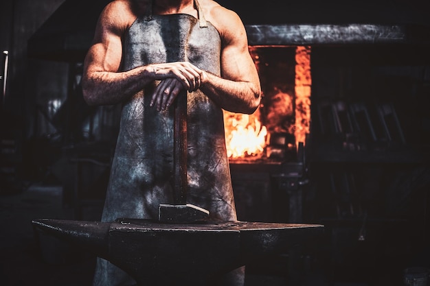 Herrero musculoso en delantal protector en su taller con martillo en las manos. Hay fuego en el fondo.