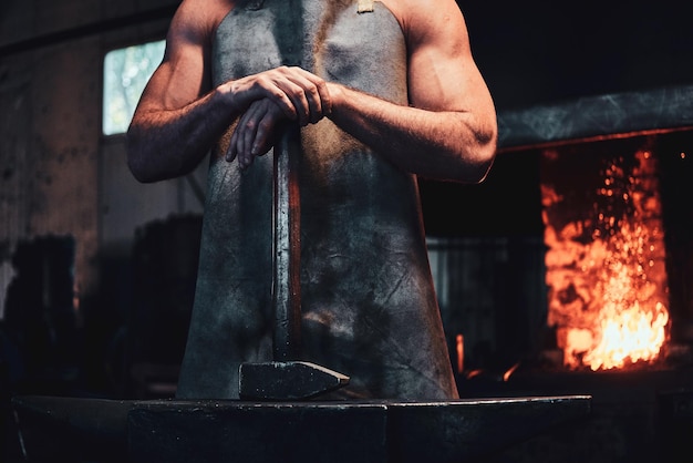 Herrero musculoso en delantal protector en su taller con martillo en las manos. Hay fuego en el fondo.