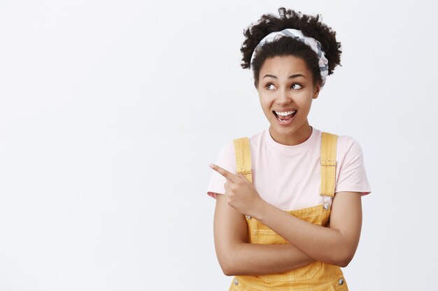 Hey Mira. Intrigada y feliz hermosa estudiante africana con cabello rizado en diadema y peto amarillo, mirando y apuntando a la izquierda con una amplia sonrisa, haciendo preguntas sobre aves interesantes