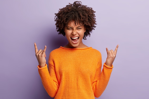 Hey, vamos a rockear esta fiesta. Emocional alegre mujer de piel oscura levanta la mano y muestra un gesto de rock n roll, exclama de alegría, disfruta de su música favorita a todo volumen, aislada contra la pared púrpura