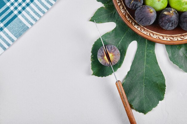 Higos verdes y negros en un plato de cerámica con un cuchillo y una hoja. Foto de alta calidad