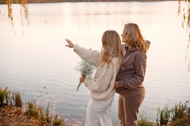 Hija adulta abraza a su madre embarazada en un día soleado de otoño en la naturaleza Mujeres rubias de pie en el parque cerca del lago Mujeres vestidas de color beige