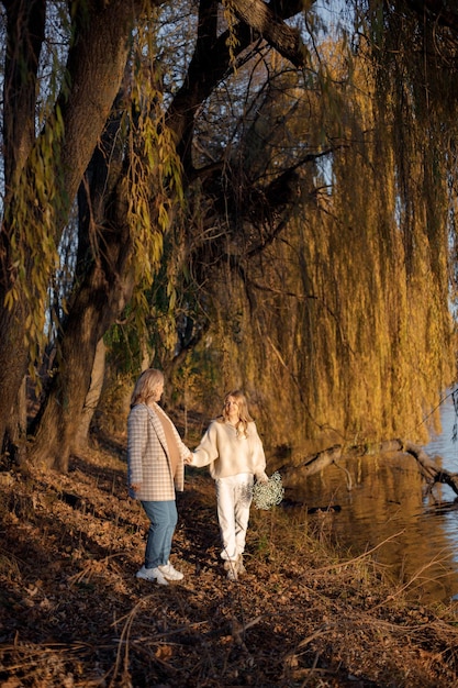 Hija adulta cogida de la mano con su madre embarazada en un soleado día de otoño en la naturaleza Mujer rubia sosteniendo flores en la mano Mujeres vestidas de color beige