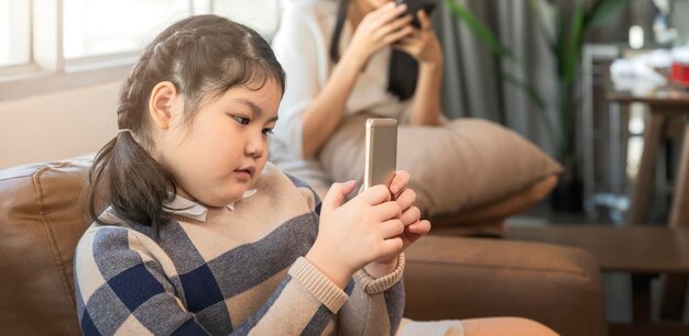 La hija asiática se concentra en el juego de teléfono inteligente de uso manual en línea jugando con una cuarentena alegre y divertida, quédese en casa en el fondo interior de la sala de estar