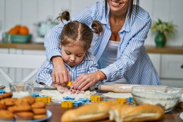 Hija ayudando a su madre a cocinar galletas durante las vacaciones. Se enrollan y cortan la masa en la cocina.