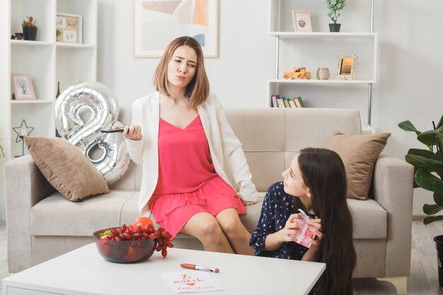 Hija contenta sosteniendo presente mirando a la madre disgustada sentada en el sofá en el día de la mujer feliz en la sala de estar