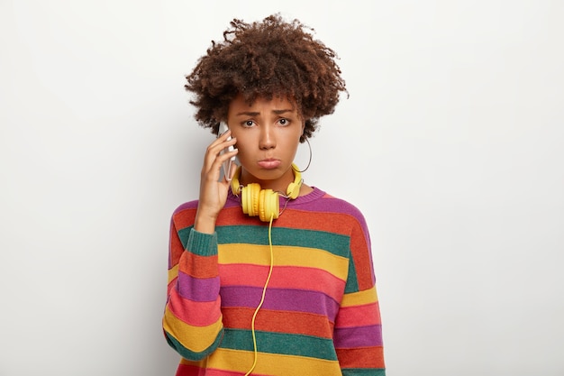 Hija descontento niña afroamericana tiene una conversación desagradable a través de un teléfono inteligente, usa un jersey de rayas casuales de colores, usa auriculares amarillos, no está satisfecha con algo