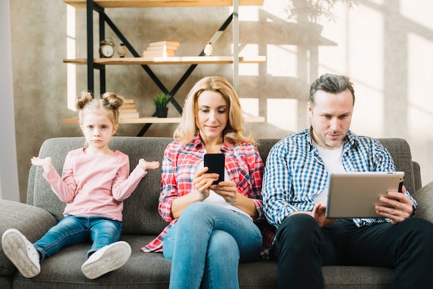 Hija enojada que se sienta en el sofá con su madre y padre que usa la tableta digital; celular en casa