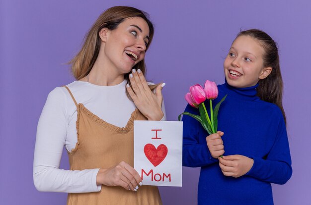 Hija feliz dando tarjetas de felicitación y flores de tulipanes para su madre sorprendida y sonriente celebrando el día internacional de la mujer de pie sobre una pared púrpura