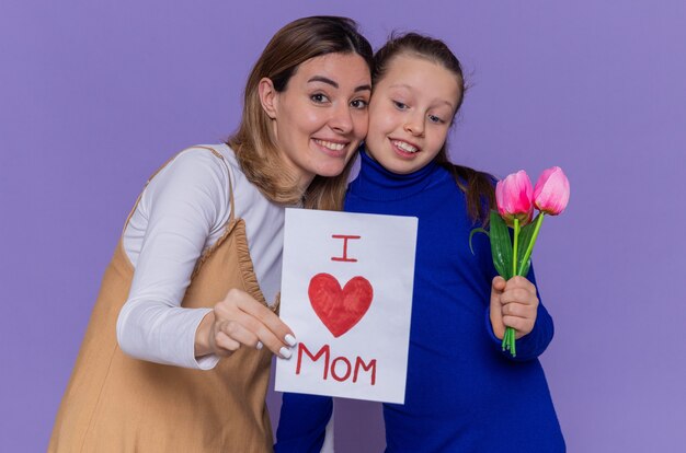 Hija feliz dando tarjetas de felicitación y flores de tulipanes para su madre sorprendida y sonriente celebrando el día de la madre de pie sobre la pared púrpura