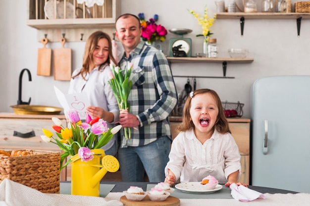 Hija graciosa haciendo cupcake cerca de padres con tarjeta de felicitación