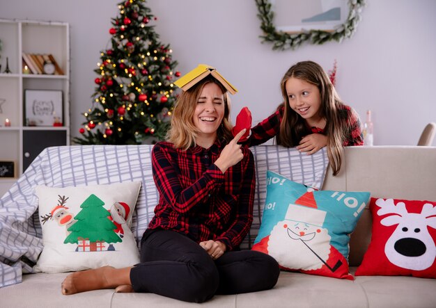 Hija juega con medias de pie detrás de su madre sosteniendo un libro en la cabeza sentado en el sofá y disfrutando de la Navidad en casa