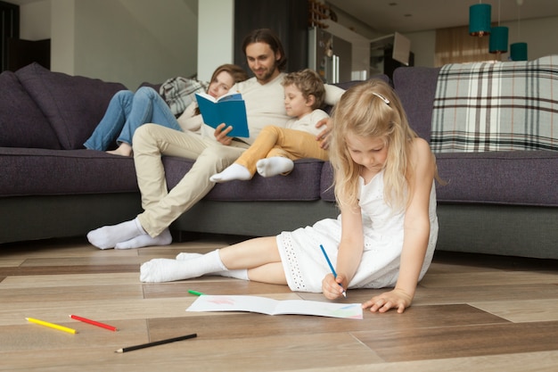 Hija jugando en el piso mientras los padres y su hijo leen un libro