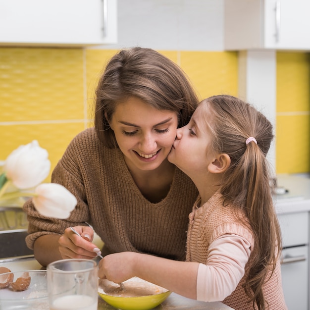 Hija linda que besa a la madre mientras que cocina en cocina