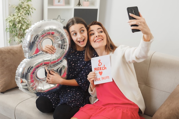 Hija y madre sorprendidas con el globo número ocho y una postal en el día de la mujer feliz sentada en el sofá toman un selfie en la sala de estar