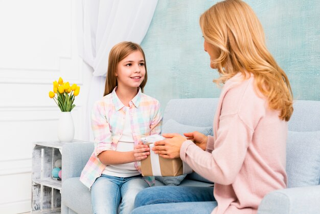 Hija que da la caja de regalo para la mamá que se sienta en el sofá