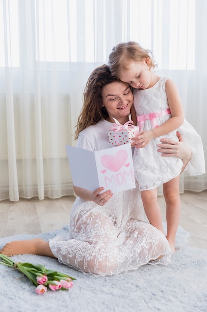 Hija que da la caja de regalo a su madre mientras que lee la tarjeta de felicitación