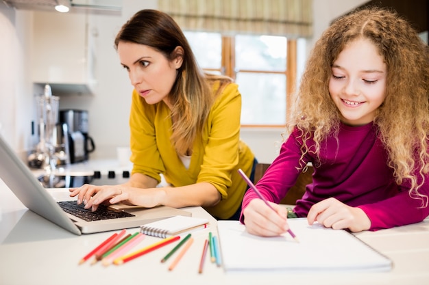 Hija que hace su preparación y su madre trabaja en la computadora portátil en kitc