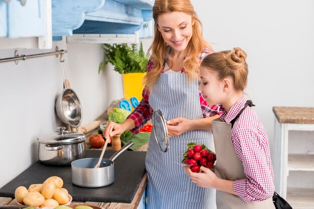 Hija que sostiene el nabo en la mano que mira a su madre que prepara la comida en la cocina