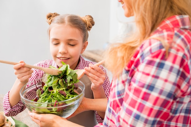 Hija que toma el olor de la ensalada de hojas frescas en el tazón que tiene su madre.