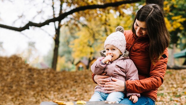 Hija sentada en las rodillas de la madre y comiendo galletas en el parque de otoño