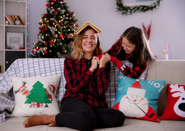 Hija sonriente juega con su madre sosteniendo el libro en la cabeza sentado en el sofá y disfrutando de la Navidad en casa