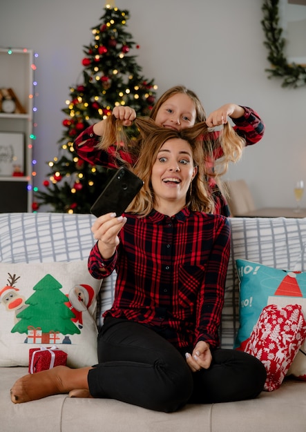 Hija sonriente levanta el cabello de su madre sosteniendo el teléfono sentado en el sofá y disfrutando de la Navidad en casa