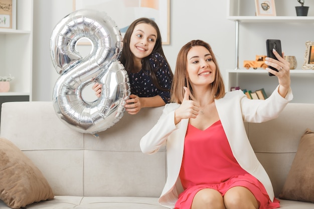 Hija sonriente de pie detrás del sofá sosteniendo la madre del globo número ocho sentada en el sofá tomar un selfie en el día de la mujer feliz en la sala de estar