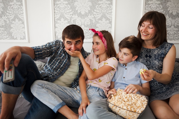 Hija sonriente sentada en el sofá con la familia y alimentando palomitas a su padre