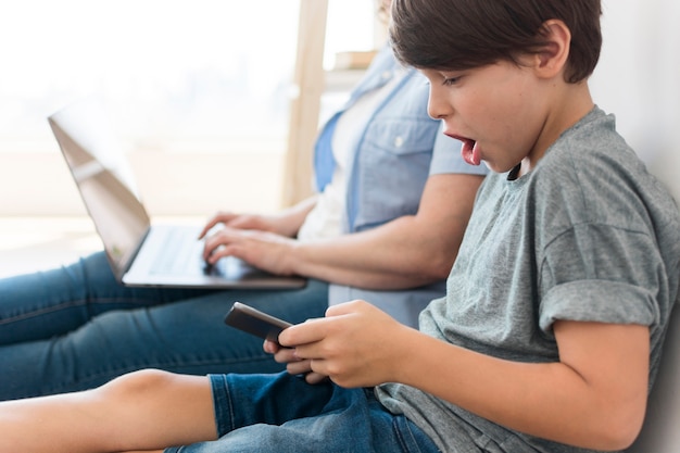 Hijo jugando en el teléfono inteligente junto a mamá