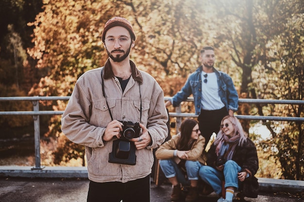 Un hipster atractivo con gafas posa para un fotógrafo con una cámara fotográfica mientras sus amigos descansan en el fondo.