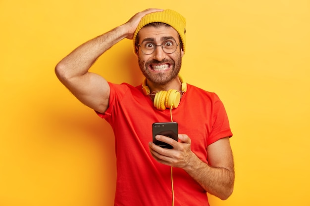 Hipster avergonzado aprieta los dientes, mira nerviosamente, no puede descargar la aplicación necesaria en un teléfono inteligente, tiene auriculares alrededor del cuello, se viste de manera informal