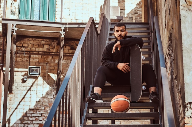 Un hipster barbudo de piel oscura vestido con una capucha negra y pantalones cortos deportivos sostiene una patineta mientras se sienta en las escaleras de un gueto.