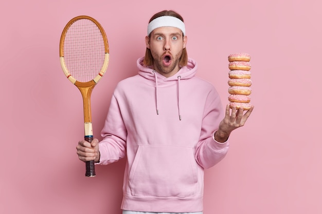 Hipster barbudo sorprendido viste atuendo deportivo sostiene raqueta de tenis y montón de rosquillas dulces miradas sorprendentemente con la boca ampliamente abierta tiene ocio activo.