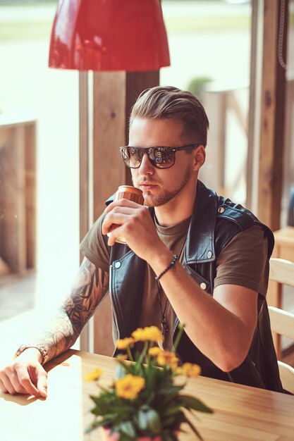 Hipster con un elegante corte de pelo y barba se sienta en una mesa en un café al borde de la carretera, bebe un refresco en un día caluroso.