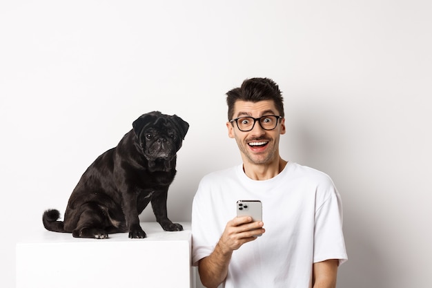 Hipster joven alegre mirando a cámara, sentado con lindo perro pug negro y usando teléfono móvil, de pie sobre fondo blanco.