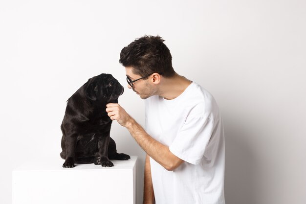 Hipster joven guapo con gafas, acaricia a su perro, mirándose, de pie sobre fondo blanco.