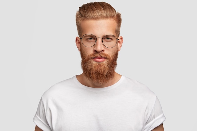 Hipster joven guapo tiene barba y bigote gruesos de jengibre, corte de pelo moderno, te mira seriamente directamente