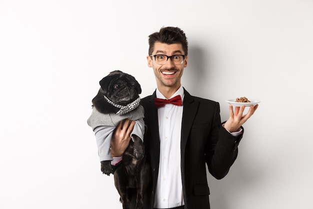 Hipster joven guapo en traje y gafas con lindo pug negro y comida para mascotas en un plato, de pie sobre fondo blanco.