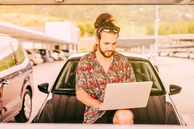 Hipster con laptop sentado en el capó del coche