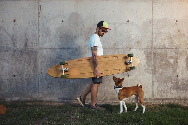 Hipster longboarder y un joven perro basenji marrón y blanco mirando el uno al otro junto a un muro de hormigón gris