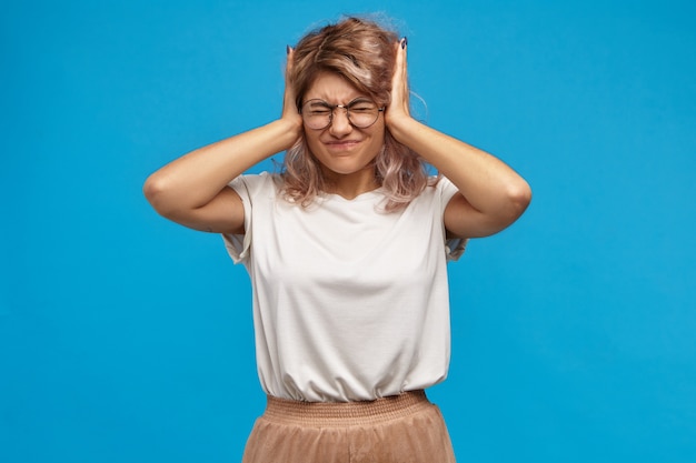 Hipster mujer enojada irritada en gafas de moda cubriendo las orejas con las manos y atornillando los ojos molestos con un sonido chirriante o música alta. Una adolescente frustrada no puede soportar que sus padres peleen