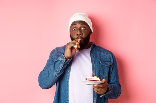 Hipster negro feliz celebrando el cumpleaños, soplando el silbato de la fiesta, sosteniendo el pastel de cumpleaños con velas, de pie sobre fondo rosa.