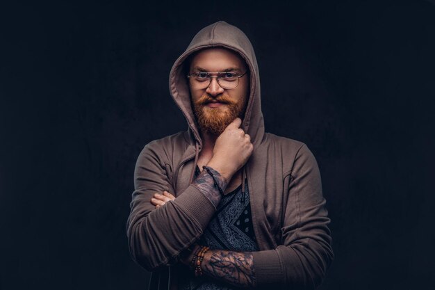 Hipster pelirrojo pensativo con barba completa y anteojos vestidos con capucha y camiseta posa con la mano en la barbilla en un estudio. Aislado en un fondo oscuro.